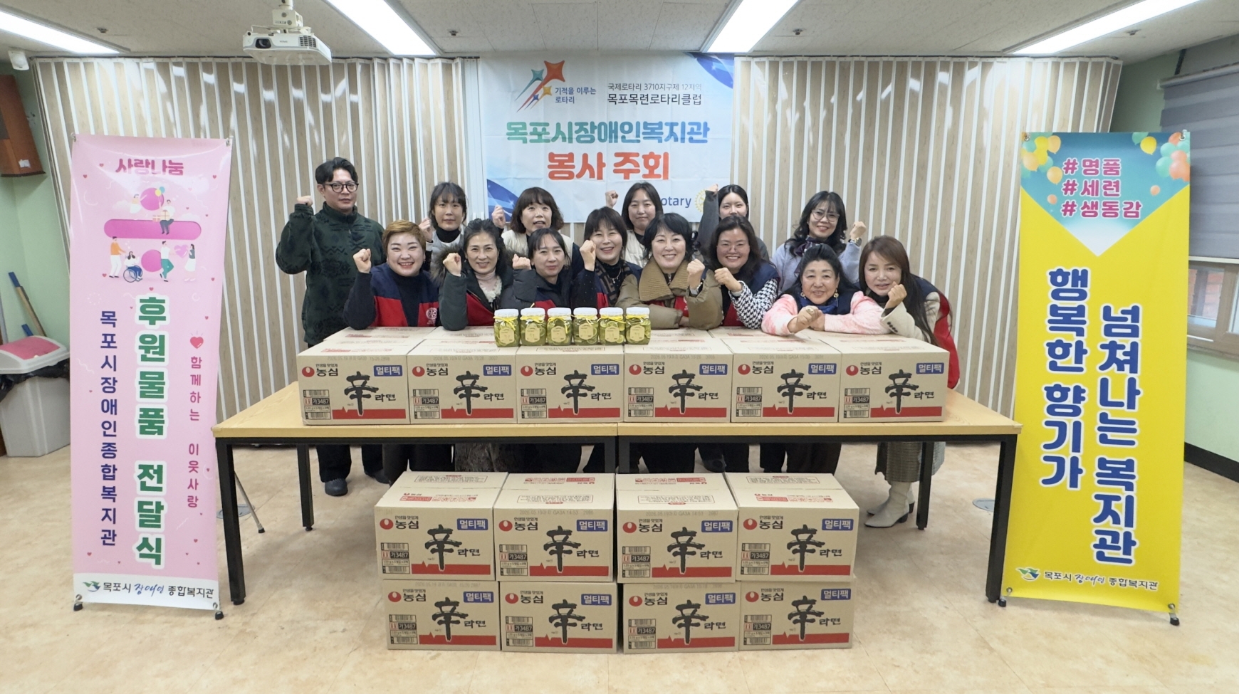 목포목련로타리클럽 회원들이 목포시장애인종합복지관에 라면을 후원하고 봉사 주회를 기념하는 촬영을 하고 있다. 테이블 위와 아래에는 농심 신라면 박스가 가득 쌓여 있고, 테이블 중앙 라면 박스 위에는 직접 담근 것으로 보이는 수제청 병들이 놓여 있다. 뒤편 벽면에는 '기적을 이루는 로타리, 목포시장애인복지관 봉사 주회, 목포목련로타리클럽'이라고 적힌 대형 현수막이 걸려 있다. 조끼를 입은 회원들을 포함한 13명의 참석자가 주먹을 쥐고 밝은 표정으로 파이팅을 외치고 있다.